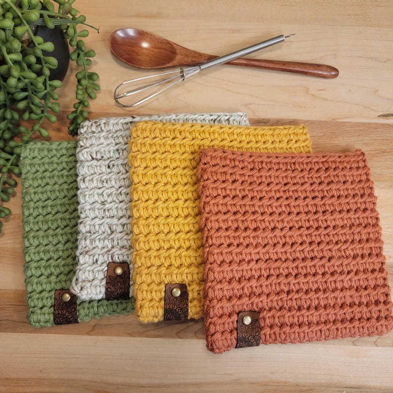 May include: A collection of four square, crocheted washcloths in shades of green, gray, yellow, and orange. Each cloth has a leather tag with a decorative pattern. A wooden spoon and whisk rest above the cloths.