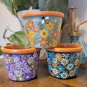 May include: Three small, hand-painted ceramic flower pots. The top pot is teal with orange, yellow, and white flowers and a bee. The other two pots have purple and blue floral designs, respectively. Each pot has an orange rim.