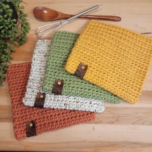 May include: A collection of handmade crochet square pot holders in shades of rust, cream, green, and yellow. Each pot holder has a decorative leather tag with a metal rivet. A wooden spoon and whisk are in the background.
