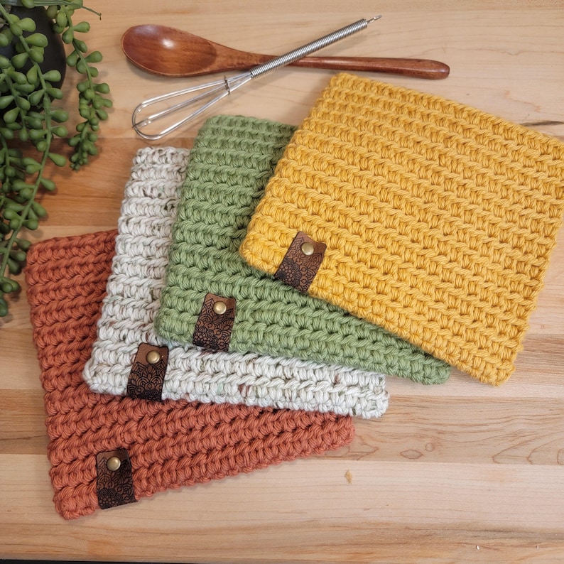 May include: A collection of handmade crochet square pot holders in shades of rust, cream, green, and yellow. Each pot holder has a decorative leather tag with a metal rivet. A wooden spoon and whisk are in the background.