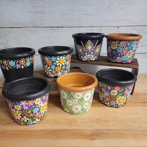 May include: A collection of seven small, hand-painted flower pots. The pots are black, brown, and tan, with colorful floral designs. Some pots feature daisy patterns, while others have mandala-style dot art. The pots are displayed on a wooden surface.