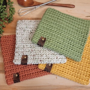 May include: Four handmade crochet pot holders in shades of orange, cream, green, and yellow. Each square pot holder has a leather tag with a brass button. Kitchen utensils, including a wooden spoon and whisk, are in the background.
