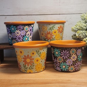May include: Four small terracotta pots with orange rims. Two pots feature a dense pattern of purple, blue, and cream daisies on a black background. The other two pots display orange and yellow flowers with green leaves on a light green background.