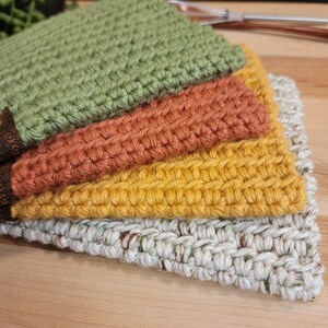 May include: A stack of four handmade crochet washcloths in various colors. The colors include green, orange, yellow, and a speckled off-white. The washcloths are made with a textured stitch and are stacked on top of each other.