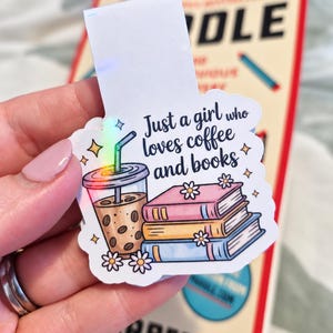 Peut inclure: Un marque-page magnétique avec un autocollant représentant un gobelet de café glacé et une pile de trois livres. Le texte indique, Just a girl who loves coffee and books. Le motif inclut des petites fleurs blanches, des étoiles jaunes et une paille holographique.
