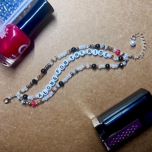 May include: A beaded bracelet with the words "ALONG FOR THE RIDE" spelled out in white letter beads. The bracelet is made with white, black, and clear beads. A red nail polish bottle and a tube of lipstick are in the background.