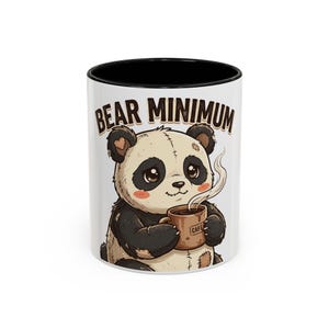 May include: White ceramic mug with a black interior featuring a cartoon panda holding a coffee cup. The panda has black and white fur and the text "BEAR MINIMUM" is above the panda.
