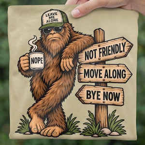 May include: A beige t-shirt featuring a brown Bigfoot wearing sunglasses and a green cap that says LEAVE ME ALONE. Bigfoot holds a mug labeled NOPE and leans against a wooden signpost with arrows reading NOT FRIENDLY, MOVE ALONG, and BYE NOW.
