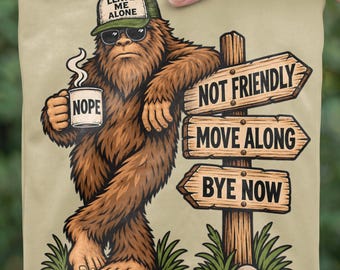 Pode incluir: Uma t-shirt bege com um Bigfoot castanho a usar óculos de sol e um boné verde com o texto LEAVE ME ALONE. O Bigfoot segura uma caneca com a palavra NOPE e apoia-se num poste de madeira com sinais que dizem NOT FRIENDLY, MOVE ALONG, BYE NOW.