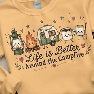 May include: A light yellow sweatshirt featuring a graphic print of a lantern, a campfire, a camper trailer, a coffee mug, and a marshmallow with faces. The text reads Life is Better Around the Campfire, surrounded by small stars, hearts, and leaves.