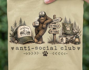 Pode incluir: Uma t-shirt bege com o gráfico de um Bigfoot castanho com um boné que diz LEAVE ME ALONE, segurando uma caneca que diz NOT TODAY. Ao lado, um poste com placas que dizem NO PEOPLE, NO PROBLEM, NO THANKS, uma cadeira e uma lanterna.