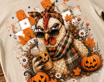 Puede incluir: Una camiseta beige con el gráfico de una gallina que lleva un pañuelo a cuadros, gafas de sol y una bufanda a juego. La gallina sostiene un vaso de café helado con una cara de calabaza. Dos calabazas y hojas de otoño rodean a la gallina.