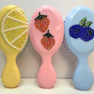 Puede incluir: Tres cepillos de pelo ovalados en amarillo pastel, rosa y azul claro. El cepillo amarillo presenta un diseño de rodaja de limón hecho de cristales amarillos y blancos. El rosa tiene dos diseños de fresas y el azul un diseño de arándanos.