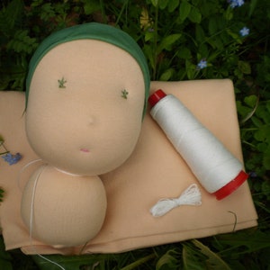 Könnte beinhalten: Eine handgefertigte Puppe mit einem grünen Stoffhut, einem hellpfirsichfarbenen Körper und einer Spule weißen Fadens mit roter Kappe. Die Puppe liegt auf einem hellpfirsichfarbenen Stoff.