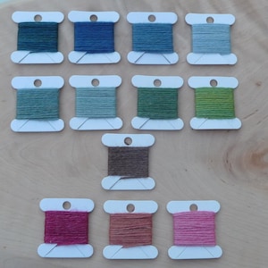 Könnte beinhalten: Eine Sammlung von 13 Spulen Stickgarn in verschiedenen Grüntönen, Braun, Rosa und Blau. Die Spulen sind in drei Reihen auf einer Holzoberfläche angeordnet.