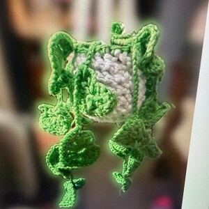 May include: A handmade crochet frog-shaped item, in white and green. The frog's body is white, with green crochet details forming the eyes, legs, and decorative elements. The item appears to be a decorative piece or a small container.