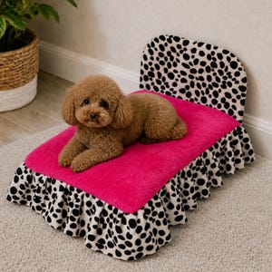Puede incluir: Un perro pequeño marrón descansa sobre una cama rosa para mascotas con cabecero y falda con estampado de lunares blancos y negros. La cama está colocada sobre una alfombra de color neutro, con una planta y cestas en el fondo.