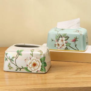 Könnte beinhalten: Zwei rechteckige Taschentuchboxen aus Keramik. Eine Box ist perlmuttweiß mit einem Blumenmuster aus weißen Blüten und grünen Blättern. Die andere Box ist hellblau mit einem ähnlichen Blumenmuster und enthält weiße Taschentücher.