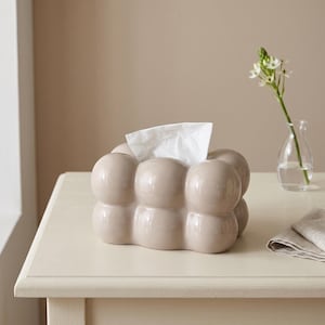 May include: A beige ceramic tissue box cover featuring a rounded, bubble-like exterior texture. The rectangular container has a central opening at the top from which a single white tissue is pulled upward, displaying a smooth, glossy finish.