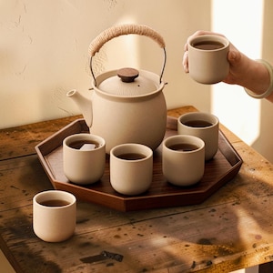 May include: A beige ceramic tea set comprising a teapot, six cups, and a wooden tray. The teapot features a woven handle and a wooden lid. The cups are filled with tea. The set is displayed on a wooden table.