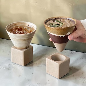 May include: Two ceramic cone-shaped cups with drinks and ice, one resting on a square base. The other cup is held by a person. The cups have brown, beige, and white tones. One cup has a sprig of rosemary.