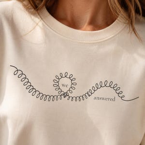Könnte beinhalten: Ein cremefarbenes Sweatshirt mit Rundhalsausschnitt und einer schwarzen Strichgrafik einer durchgehenden Spiralfeder. Das Wort we ist in einer kreisförmigen Spirale gedruckt, und das Wort answered steht in schwarzer Schrift unter der Spirale.