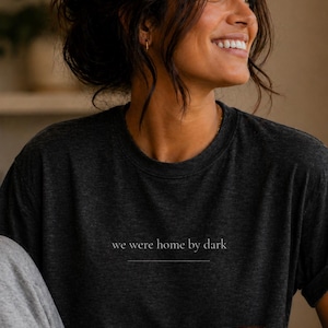 以下が含まれることがあります： 「we were home by dark」のテキストがシンプルなフォントでプリントされたダークグレーのTシャツ。柔らかい素材でできており、クルーネックのデザインです。白いマグカップが手に持たれています。