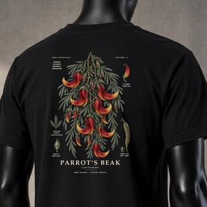 Könnte beinhalten: Ein schwarzes Kurzarm-T-Shirt mit einer botanischen Illustration auf der Rückseite. Die Grafik zeigt ein Büschel roter und gelber Papageienschnabel-Blüten mit grünem Blattwerk, ergänzt durch kleine Diagramme und den Text Parrot's Beak.