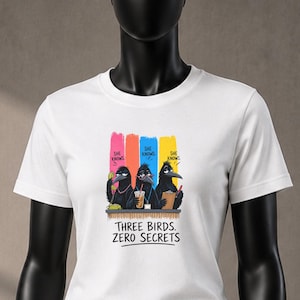 May include: A white short-sleeved t-shirt featuring a graphic of three black birds with colorful vertical stripes behind them. The text above the birds reads "SHE KNOWS" and the text below reads "THREE BIRDS. ZERO SECRETS" in black lettering.