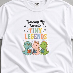 May include: White t-shirt with the text "Teaching My Favorite Tiny Legends" and a graphic of three cartoon dinosaurs reading books. The dinosaurs are blue, pink, and green, with colorful accents.