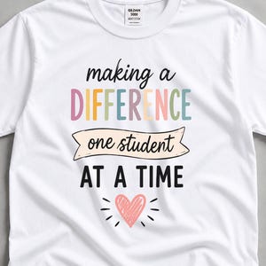 Può includere: T-shirt bianco con la scritta "making a DIFFERENCE one student AT A TIME" in lettere colorate. Un cuore rosa con linee radianti è sotto il testo. La maglietta è una Gildan 5000.