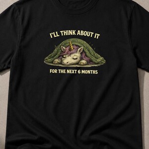 Könnte beinhalten: Ein schwarzes Kurzarm-T-Shirt mit einer Grafik eines Einhorns mit violetter Mähne, das unter einer grünen Decke schläft. Der Text über dem Einhorn lautet I'LL THINK ABOUT IT und der Text unter dem Einhorn lautet FOR THE NEXT 6 MONTHS.
