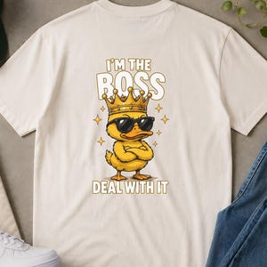 Può includere: T-shirt color crema con un'anatra dei cartoni animati con occhiali da sole e una corona, con la scritta "I'M THE BOSS DEAL WITH IT". L'immagine include anche un berretto da baseball verde, occhiali da sole, scarpe da ginnastica bianche e jeans blu.