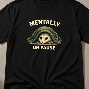 Puede incluir: Una camiseta negra de manga corta con un gráfico de un gato pequeño asomándose desde debajo de una manta verde oscuro. El texto MENTALLY está impreso en color crema sobre el gato, y el texto ON PAUSE está impreso en color crema debajo.