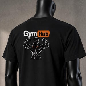 Könnte beinhalten: Ein schwarzes T-Shirt mit kurzen Ärmeln und einem weißen Grafikdruck auf dem Rücken, der eine muskulöse Figur beim Posen zeigt. Über der Figur steht der Text Gym Hub in Weiß und Orange. Das T-Shirt wird auf einer dunklen Schaufensterpuppe präsentiert.