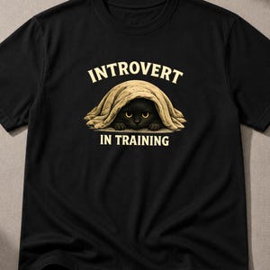 Può includere: Una t-shirt nera a maniche corte con la grafica di un gatto nero nascosto sotto una coperta beige. Sopra il gatto, il testo recita INTROVERT, e sotto il gatto, il testo recita IN TRAINING. Il gatto ha occhi gialli visibili che spuntano.