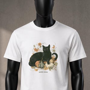 Può includere: Una t-shirt bianca a maniche corte con una grafica raffigurante un gatto nero che riposa tra zucche e foglie autunnali. Sotto l'illustrazione, il testo GOURD-GEOUS è stampato in un semplice carattere nero. La maglia ha un girocollo e orlo dritto.