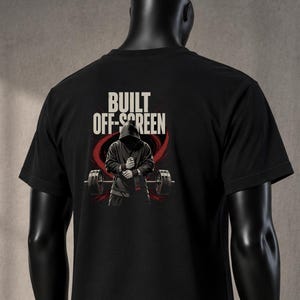 Könnte beinhalten: Ein schwarzes kurzärmeliges T-Shirt mit einem Grafikdruck auf der Rückseite. Das Motiv zeigt eine Kapuzenfigur vor einer Langhantel mit dem weißen Schriftzug BUILT OFF-SCREEN darüber. Das T-Shirt wird von einer schwarzen Schaufensterpuppe getragen.