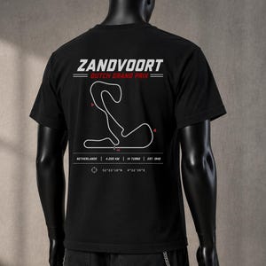 Puede incluir: Una camiseta negra con un gráfico blanco del circuito de Zandvoort en la espalda. El texto dice ZANDVOORT DUTCH GRAND PRIX, NETHERLANDS | 4.259 KM | 14 TURNS | EST. 1948, junto a las coordenadas 52°23'18"N 4°32'35"E y pequeños detalles rojos.