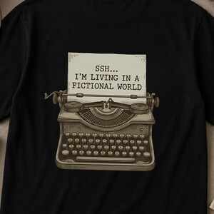 Può includere: T-shirt nero con un'illustrazione di macchina da scrivere vintage e la scritta "SSH... I'M LIVING IN A FICTIONAL WORLD" su un foglio di carta. La macchina da scrivere è nei toni del marrone e beige.