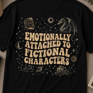 Può includere: T-shirt nera con la scritta "Emotionally Attached to Fictional Characters" in stile retrò. Il design include un drago, un libro, una luna, un sole e costellazioni color crema.