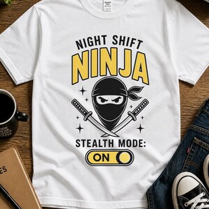 Può includere: Maglietta bianca con grafica "NIGHT SHIFT NINJA" in nero e giallo. Il design include un ninja con spade incrociate e la scritta "STEALTH MODE: ON". L'immagine mostra anche jeans, scarpe da ginnastica e una tazza di caffè.