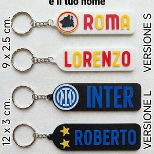 Op de afbeelding: Sleutelhangers met team- en spelersnamen. De sleutelhangers zijn wit, zwart en blauw, met teamnamen in geel en rood. Ze meten 9 x 2,5 cm en 12 x 3 cm.