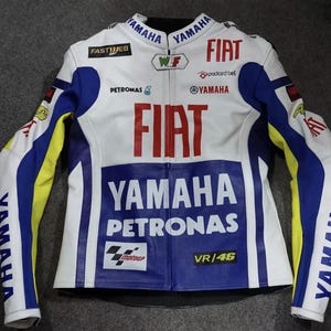 May include: A white, blue, and yellow motorcycle racing jacket featuring prominent red FIAT branding, YAMAHA and PETRONAS logos, and the text VR/46. The jacket includes various sponsor patches such as FASTWEB, WLF, and the MotoGP logo on the front.