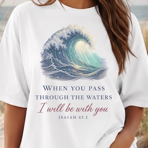 May include: A white short-sleeved t-shirt featuring a graphic of a large blue ocean wave. Below the wave, the text reads: WHEN YOU PASS THROUGH THE WATERS I will be with you ISAIAH 43 2. The shirt has a crew neckline and a relaxed fit.