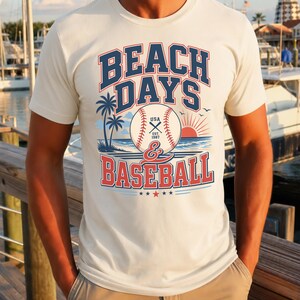 May include: A cream-colored short-sleeve t-shirt featuring a graphic print with the text BEACH DAYS & BASEBALL. The design includes a baseball, palm trees, a sunset over the ocean, and the text USA EST. 1987, rendered in navy blue and red ink.