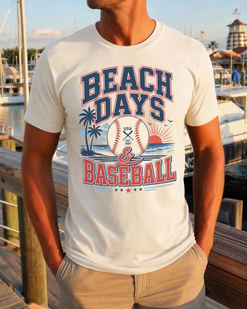 May include: A cream-colored short-sleeve t-shirt featuring a graphic print with the text BEACH DAYS & BASEBALL. The design includes a baseball, palm trees, a sunset over the ocean, and the text USA EST. 1987, rendered in navy blue and red ink.