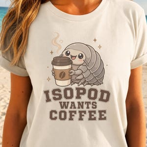 Może przedstawiać: Kremowy t-shirt z grafiką przedstawiającą kreskówkowego równonoga trzymającego kubek kawy. Tekst pod ilustracją brzmi ISOPOD WANTS COFFEE brązowymi literami blokowymi. Wzór zawiera małe akcenty w kształcie gwiazdek i parę unoszącą się z kubka.