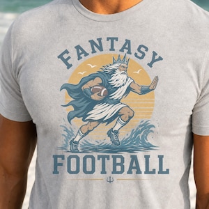 May include: A light gray t-shirt featuring a graphic of a bearded figure wearing a crown and cape, running while holding a football. The design includes the text FANTASY FOOTBALL, a yellow sun, and blue ocean waves, with a small trident icon below.