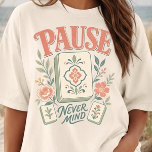 May include: An off-white short-sleeve t-shirt featuring a graphic print with the text PAUSE and NEVER MIND. The design includes three rectangular tiles with floral patterns, surrounded by green leafy branches and pink flowers in a muted color palette.